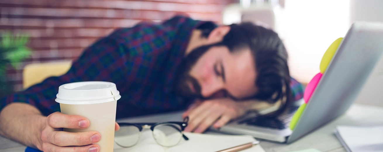Vermoeide creatieve redacteur die een wegwerpbeker vasthoudt terwijl hij op het bureau slaapt