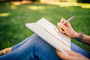 photo d'une femme assise dans l'herbe, un cahier sur les genoux et un crayon dans la main